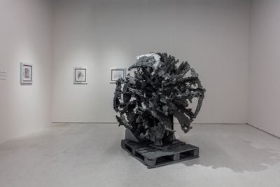 A cast zinc sculpture by Matthew Barney is spherical in structure, appearing to have parts missing from it. The sculpture is placed on a cast zinc wooden crate.