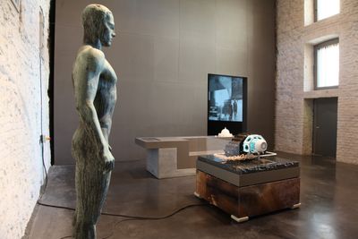 A statue of a figure stands erect in an exhibition space that also features machinic elements placed on pedestals.
