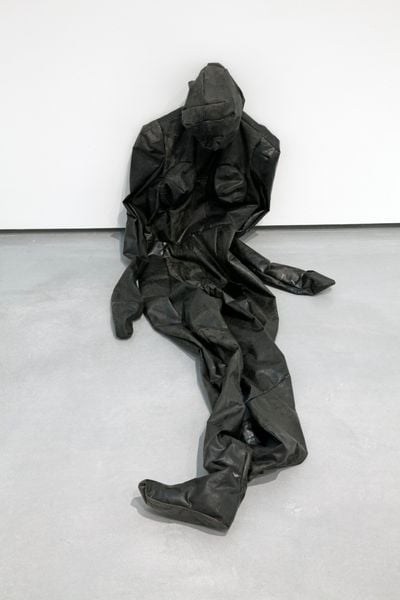 A black 'skin' in the shape of a human figure is placed slumped against a white wall in the gallery space.
