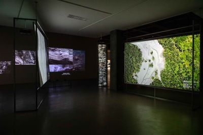 Multiple screens show aerial footage of landscapes, shown in a darkened exhibition space.