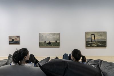 Two audience members sit on inflatable chairs in the gallery space, looking at three paintings on the gallery wall of seascapes, each featuring black geometric shapes sitting atop the waves and positioned on the horizon line.