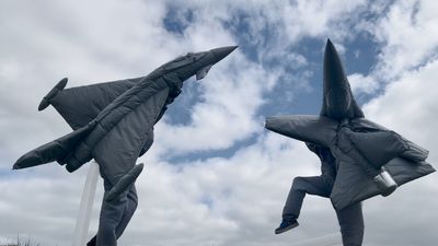 Two figures with their bodies covered in decoy airplanes dance against a cloudy sky, in Fiona Banner aka The Vanity Press's film, Pranayama Organ.