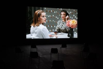 A film screen shows two women sitting at a table adorned with a flower in a vase. One holds a cigarette and looks forward, while the figure in the foreground looks across her shoulder and off to the right.