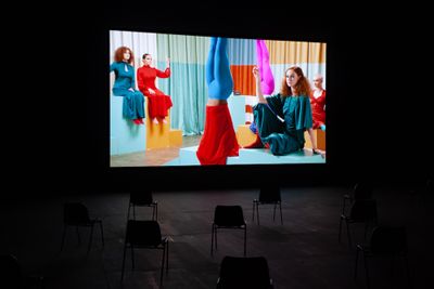 A group of women on a film set draped in colourful fabric, are also wearing colourful clothes and sit around one figure who is standing on their head. The film is shown in a darkened gallery space.