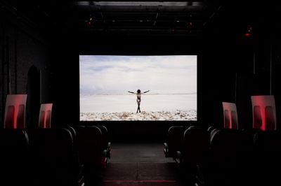 A video screen in a darkened gallery space features a figure walking towards the horizon in a barren landscape, their arms raised in the air.