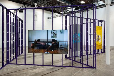 A purple metallic structure forms an installation by Martine Syms showing in a gallery space, with video screens hanging from it. One screen features a woman lying on a sofa.