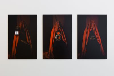 Three photographs hanging on a white wall each figure draping red fabric, from beneath which hands offer up objects such as a lamp and a mirror.