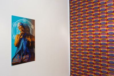 An installation photograph features patterned wallpaper pasted on a wall to the right, with a portrait of a figure hanging on the wall to the left.