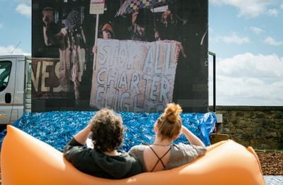 Two members of the public are sitting on an inflatable chair outdoors, viewing a screen in front of them.