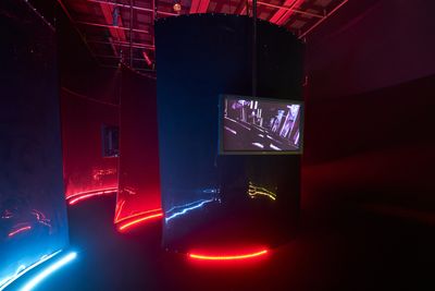 A dark room features rows of black sheets that are lit from below with neon tubing and feature a screen.