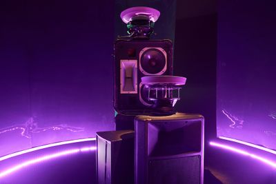A sound system sits in a purple-hued gallery space with trays placed on top of it to form a sonic art installation.