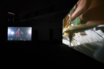 Two large screens in a darkened exhibition space show musicians playing. The right screen shows a close up of a purcussionist's hand, while the right-hand screen pictures a singer.