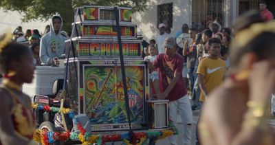 A colourful sound system is at the centre of the image, surrounded by people partying in the street.