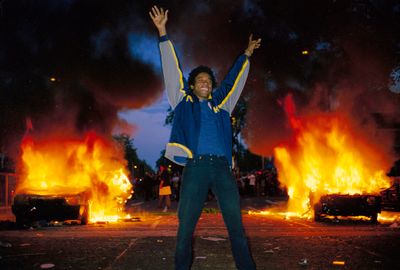 A giclée print photograph by artist Vince Peterson of a grinning man standing up in the middle of a road while holding both his arms up in celebration, with two burning cars in the background