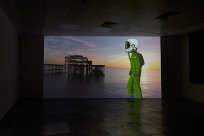 A big screen in a dark room showing an illustration of a man in a green spacesuit with white space helmet, against an image of a wharf structure at sea, a video still of Larry Achiampong's work