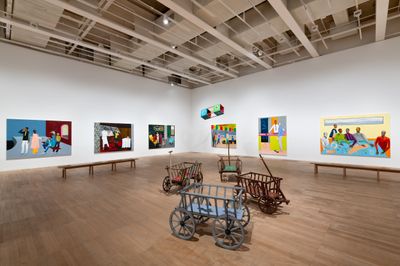 Four wooden strollers on wheels framed by large paintings in the exhibition space.  