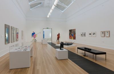 Paintings, installations, cardboard sculptures, display case, and bench inside the exhibition space. 