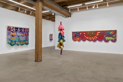 Two tapestries by Mokin Cherry hang on a wall each in a brightly lit exhibition space, foregrounded by a colourful hanging fabric sculpture. The tapestries feature surrealistic forms in different colours, while the sculpture is formed of a series of petal-shaped forms that hang off of a central column.