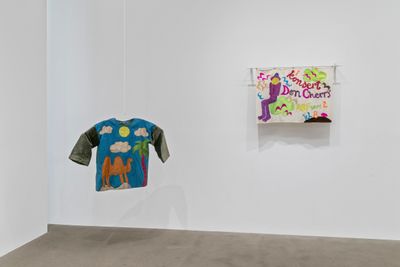 A T-shirt hangs in the corner of a white exhibition space, featuring a camel under a sound and clouds, beneath a palm tree, all rendered on a blue background. The sleeves of the T-shirt are olive green. To the right of the T-shirt, a a purple figure surrounded by fluttering birds, their face concealed by a bright yellow sun.