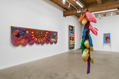 Biomorphic shapes rendered in different swatches of coloured fabric dangle off of a central column. The fabric sculpture hangs from the ceiling, surrounded by colourful, surrealistic tapestries.