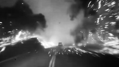 A blurred black and white film still features a car driving through what resembles a blazing forest.