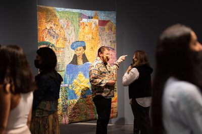 Man speaking in front of large padded canvas painting of Black woman in blue hat and coat.