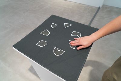 A small patch of fabric atop a pedestal is photographed from above, capturing a hand that is entering the frame to touch one of the sewn electronic segments.