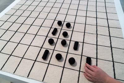 Black spherical and cylindrical shapes are placed within a grid woven in black on a white tapestry. The photograph captures a hand moving in to adjust one of the shapes on the grid.