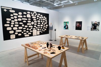 A large diagram hangs on the wall in the gallery space, overlooking two tables, each of which has a series of objects laid out across its surface.