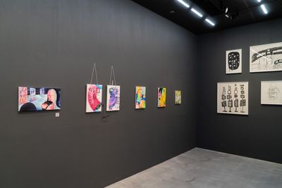 Small, colourful paintings, some abstract and others featuring figures, are hung in a line on a grey wall in the gallery space.