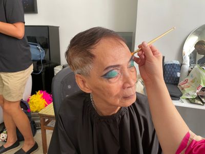 An elderly gentleman sits with his eyes closed as a hand applies exuberant eyeshadow to his face.