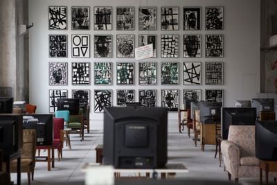 Installation showing rows of old televisions in front of chair facing 32 black and white abstract paintings.
