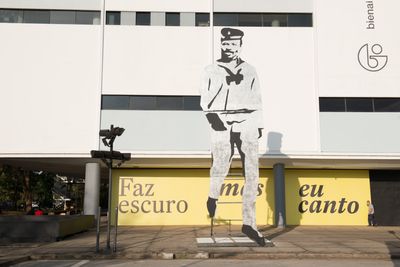 A large cut-out of a figure in uniform with a bow tie and cap by Paulo Nazareth stands outside the main exhibition hall of the Sao Paulo Bienal.