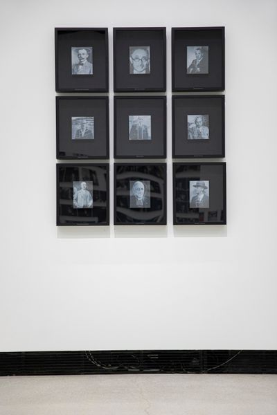 A series of 9 archival, black and white photographs of men are shown in black frames and arranged in a grid on a white gallery wall.
