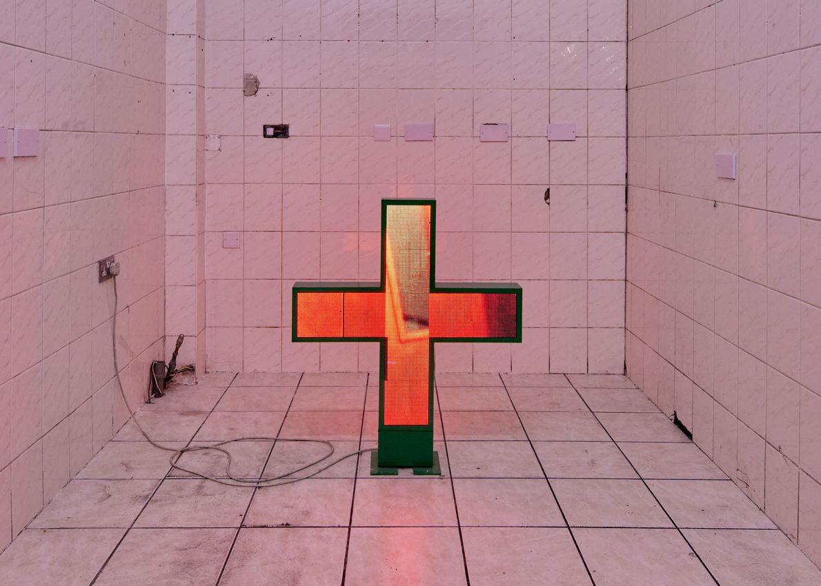 A glowing, orange, cross-shaped pharmacy sign stands on the floor in a white tiled room, exuding a minimalist, industrial feel. The space appears empty, with wires on the floor.